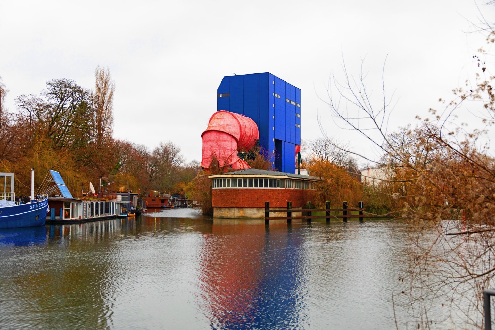 Moderne Architektur mit blauem und rotem Gebäude am Wasser, umgeben von herbstlichen Bäumen.