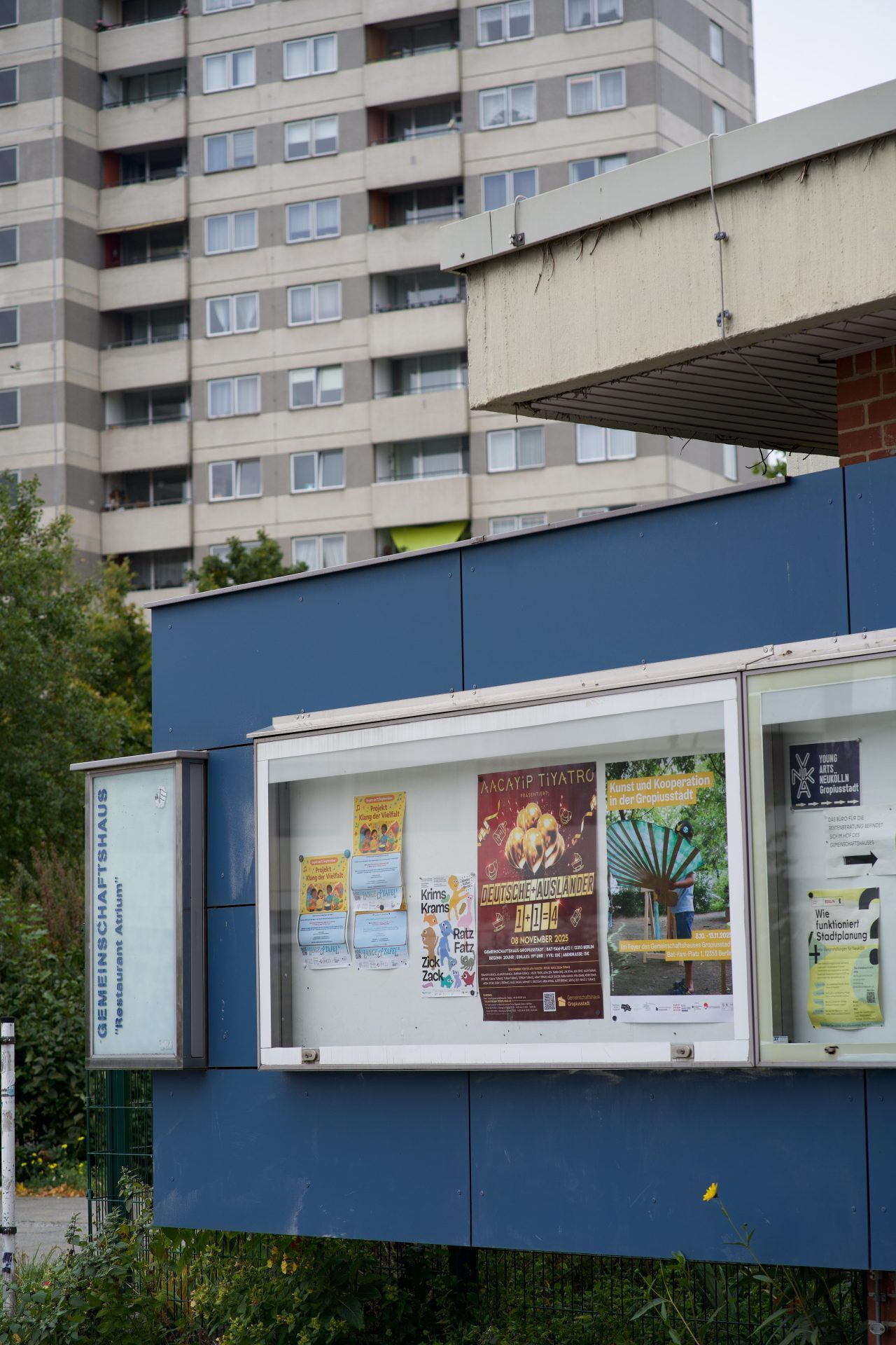 Poster wall in front of a community house with various events; a multi-story residential building in the background.