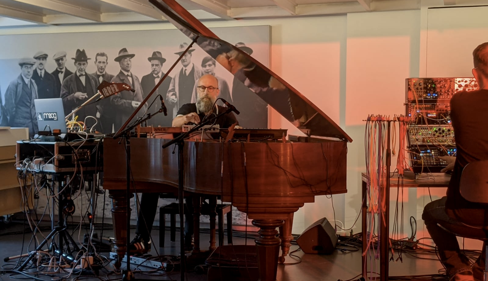 Der Musiker spielt am Klavier, umgeben von Synthesizern und Technik, während im Hintergrund historische Fotografien zu sehen sind.