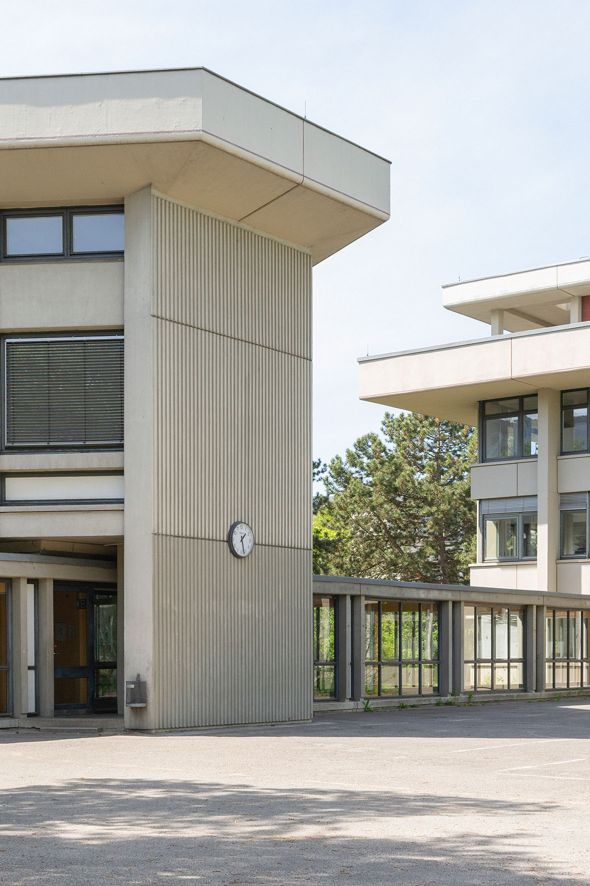 Moderne Architekturelemente eines Gebäudes mit Uhr, Fensterfronten sowie Bäumen und Parkplatz in der Umgebung.