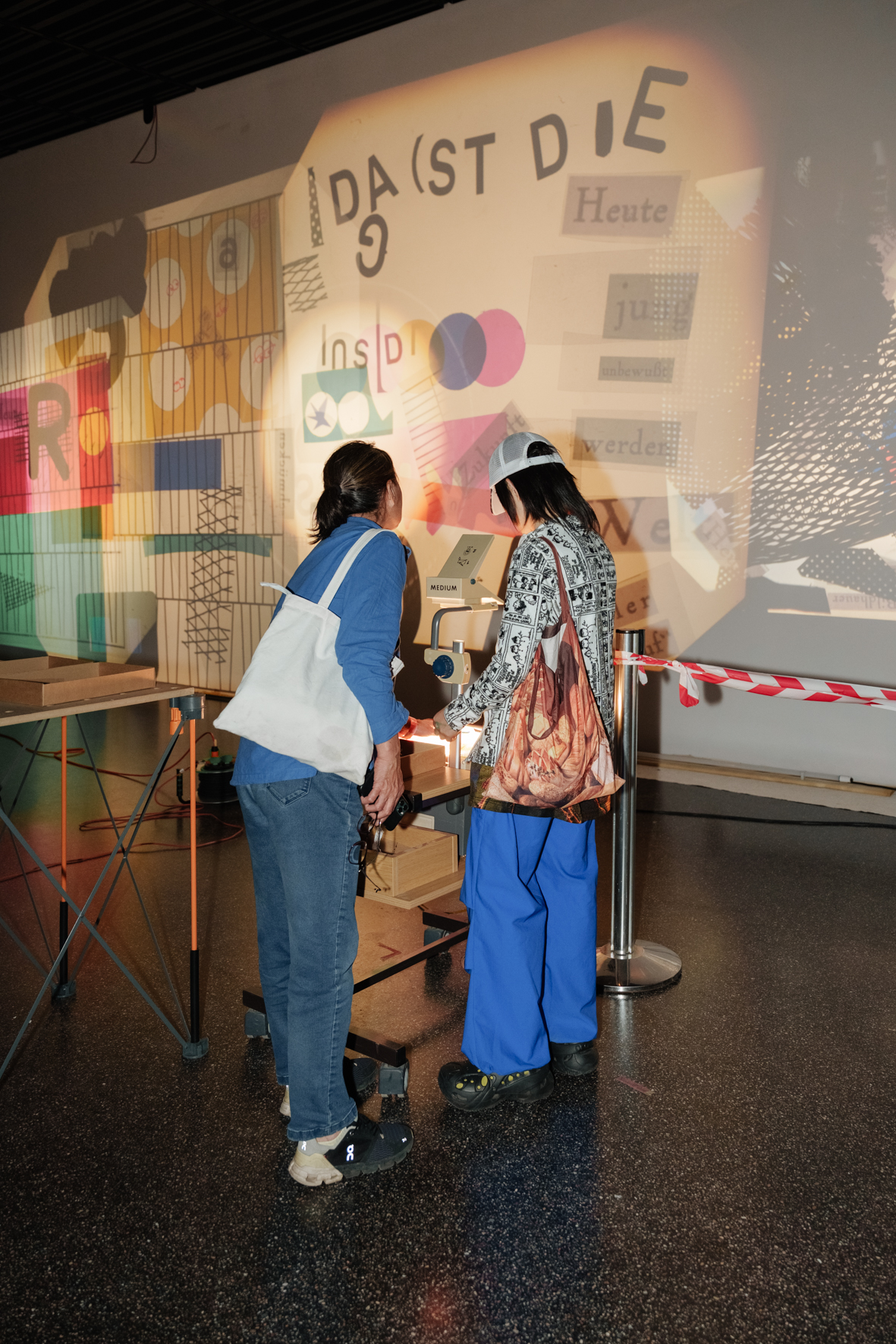Zwei Personen interagieren mit einem Ausstellungselement vor einer bunten, kreativen Wandprojektion.