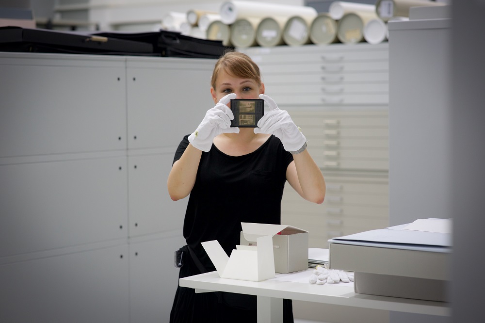 Eine Person in weißen Handschuhen zeigt ein Objekt in einer Archivanlage der Kamera.