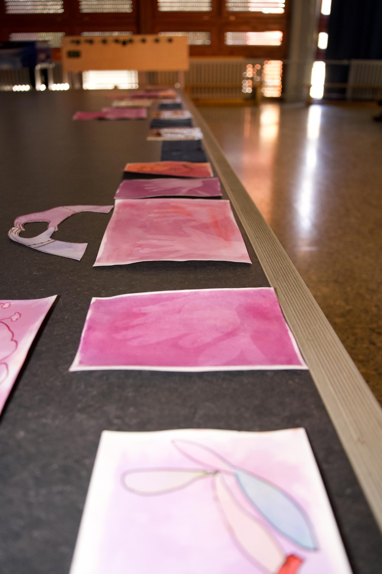 Eine Reihe von Aquarellbildern in verschiedenen Rosa- und Lilatönen liegt auf einem Tisch.