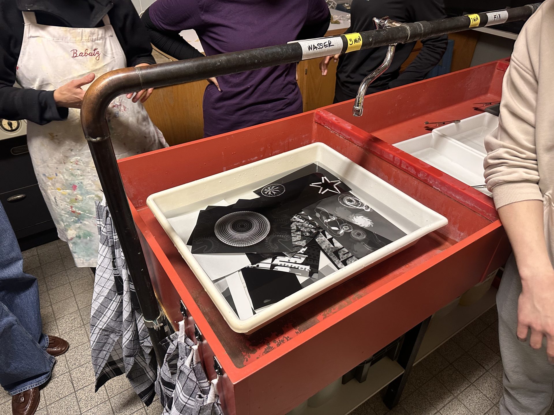 Schwarz-weiß-Fotodrucke liegen in einer Wanne mit Wasser, umgeben von Menschen in einer kreativen Umgebung.
