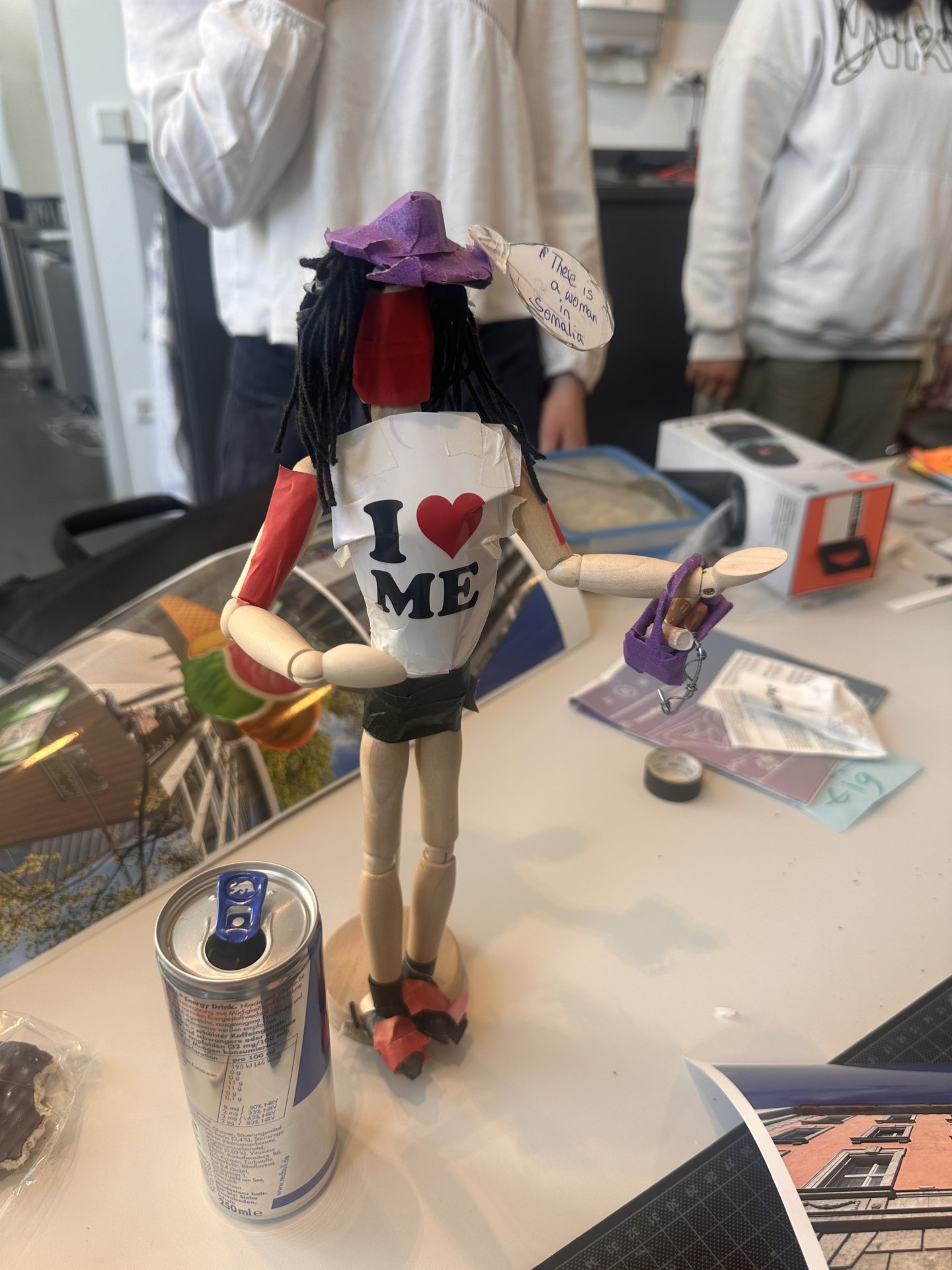 Eine handgemachte Puppe mit T-Shirt "I ❤️ ME", lila Hut und Dreadlocks steht auf einem Tisch neben einer Getränkedose.