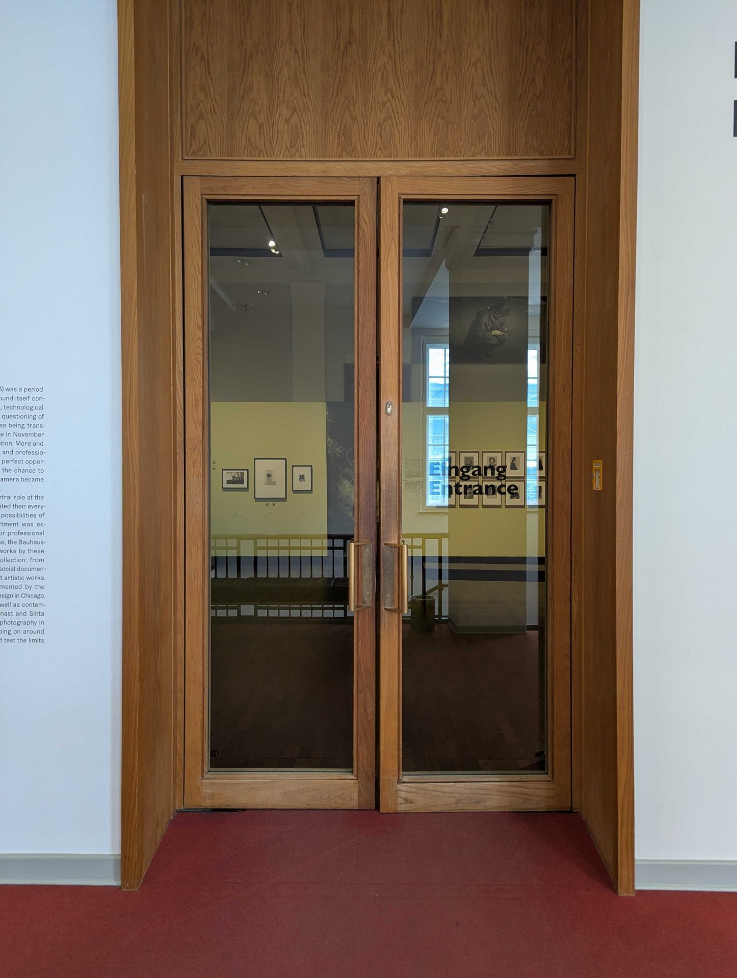 Holztüren mit Glaseinsätzen, beschriftet mit „Eingang / Entrance“, zeigen den Zugang zu einem Ausstellungsraum.