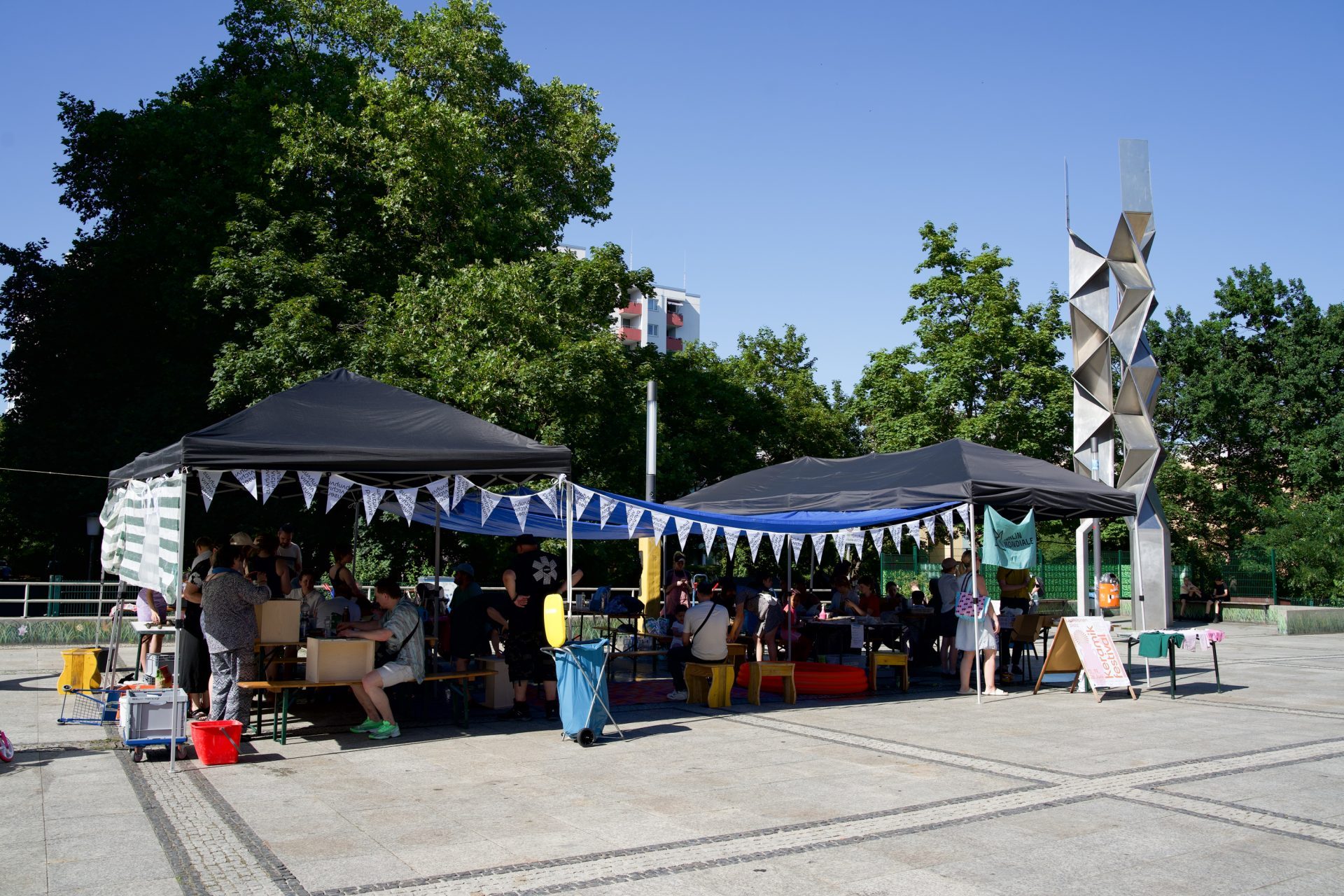 Markt mit bunten Ständen unter Zelten, Menschen interagieren, umgeben von Bäumen und einer modernen Skulptur.