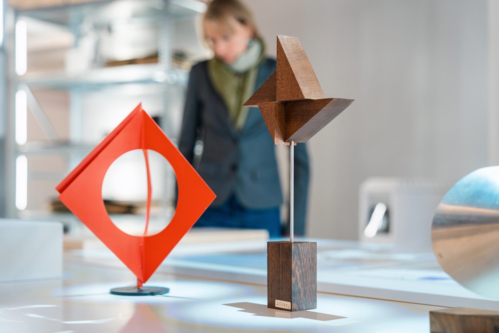 Abstracte Holz- und Metallskulpturen auf einem Tisch, im Hintergrund eine Person in geschäftlicher Kleidung.