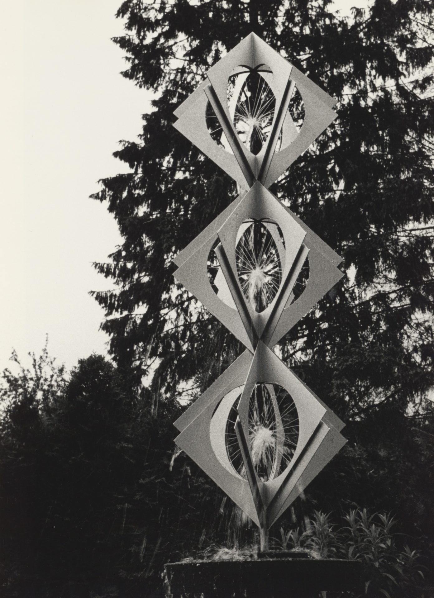 Skulptur aus Papier und Metall mit geometrischen Formen, umgeben von Bäumen, in Schwarz-Weiß-Foto.