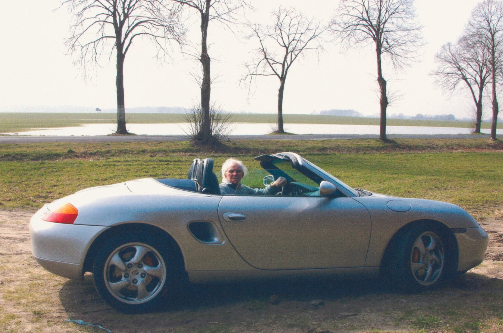 Ein älterer Mann sitzt lächelnd in einem silbernen Cabrio auf einer Wiese mit Bäumen und Wasser im Hintergrund.
