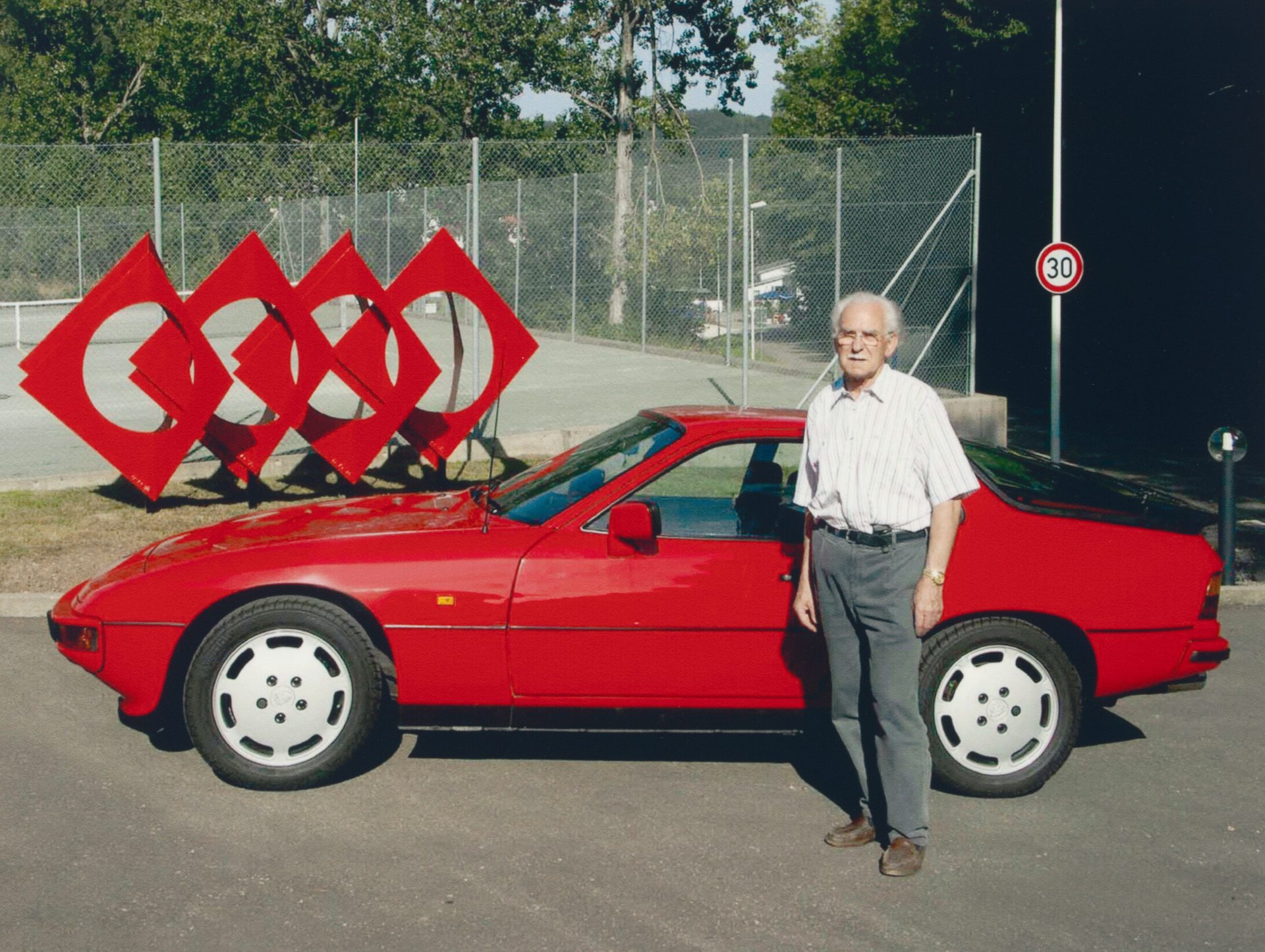 Ein älterer Mann steht neben einem roten Auto vor einer skulpturalen Installation aus roten Rhomben.