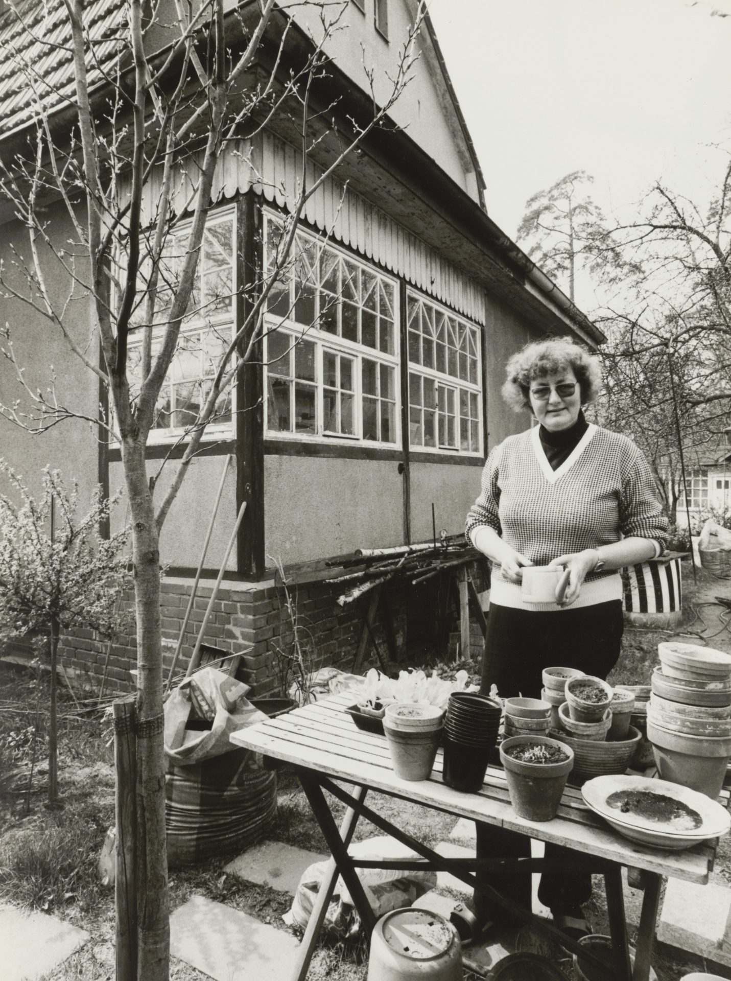 Frau mit Brille und lockigem Haar steht an einem Tisch mit Pflanzen-Töpfen vor einem Wohnhaus im Garten.