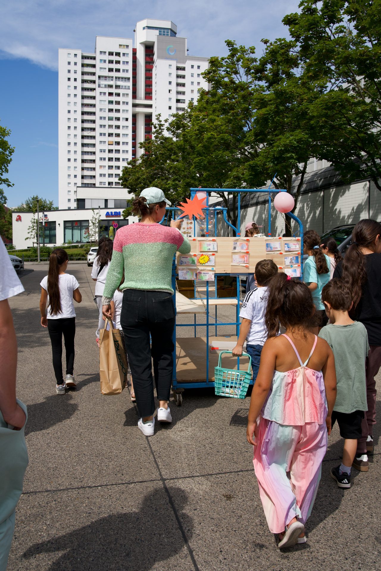 Gruppe von Kindern und Erwachsenen geht mit einem Wagen voller Waren in Richtung eines Wohngebäudes unter blauem Himmel.